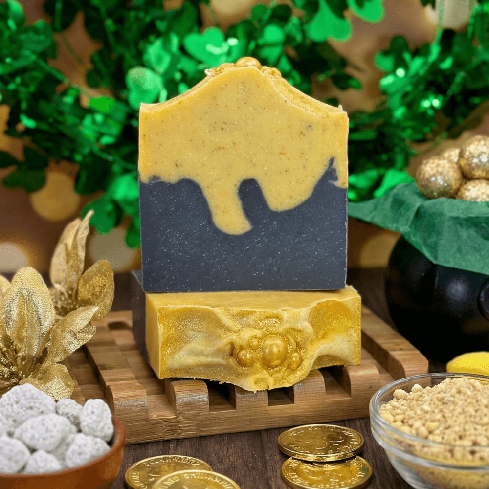 Handcrafted soap bar with gray bottom layer and yellow-gold top layer with dripping effect, decorated with gold pearls. Displayed on wooden soap dish with gold coins and green shamrock decor.