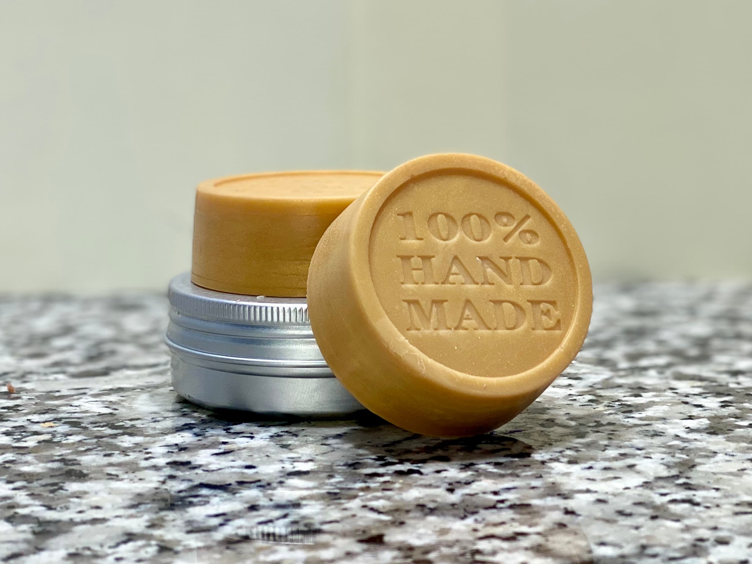 Two golden-beige circular solid lotion bars with embossed '100% HAND MADE' text, displayed with silver tin containers on a speckled granite surface against a neutral background.