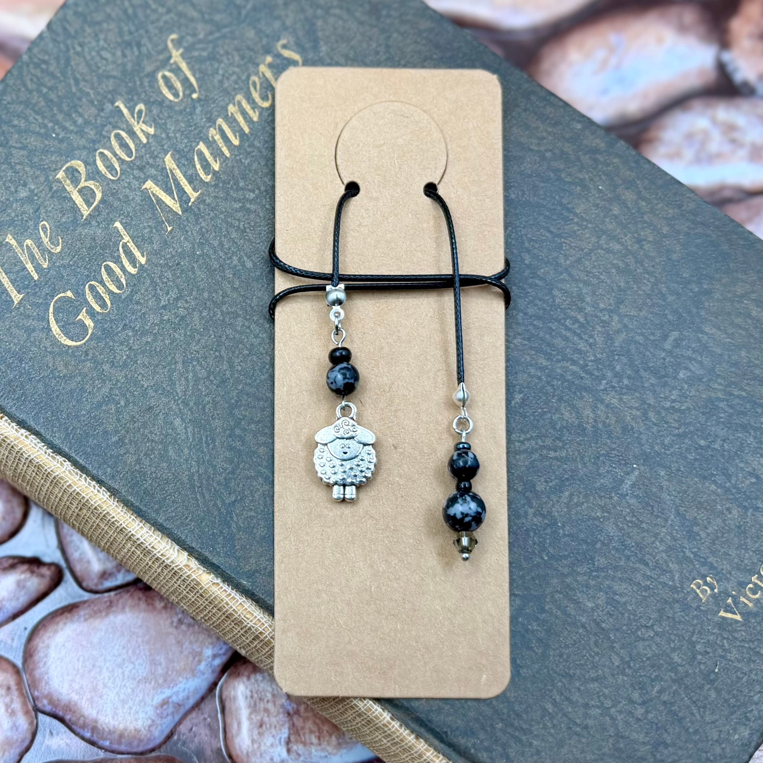 Handmade bookmark with black cord and silver sheep charm with black and gray speckled beads, displayed on a tan card atop a dark book titled 'The Book of Good Manners'.