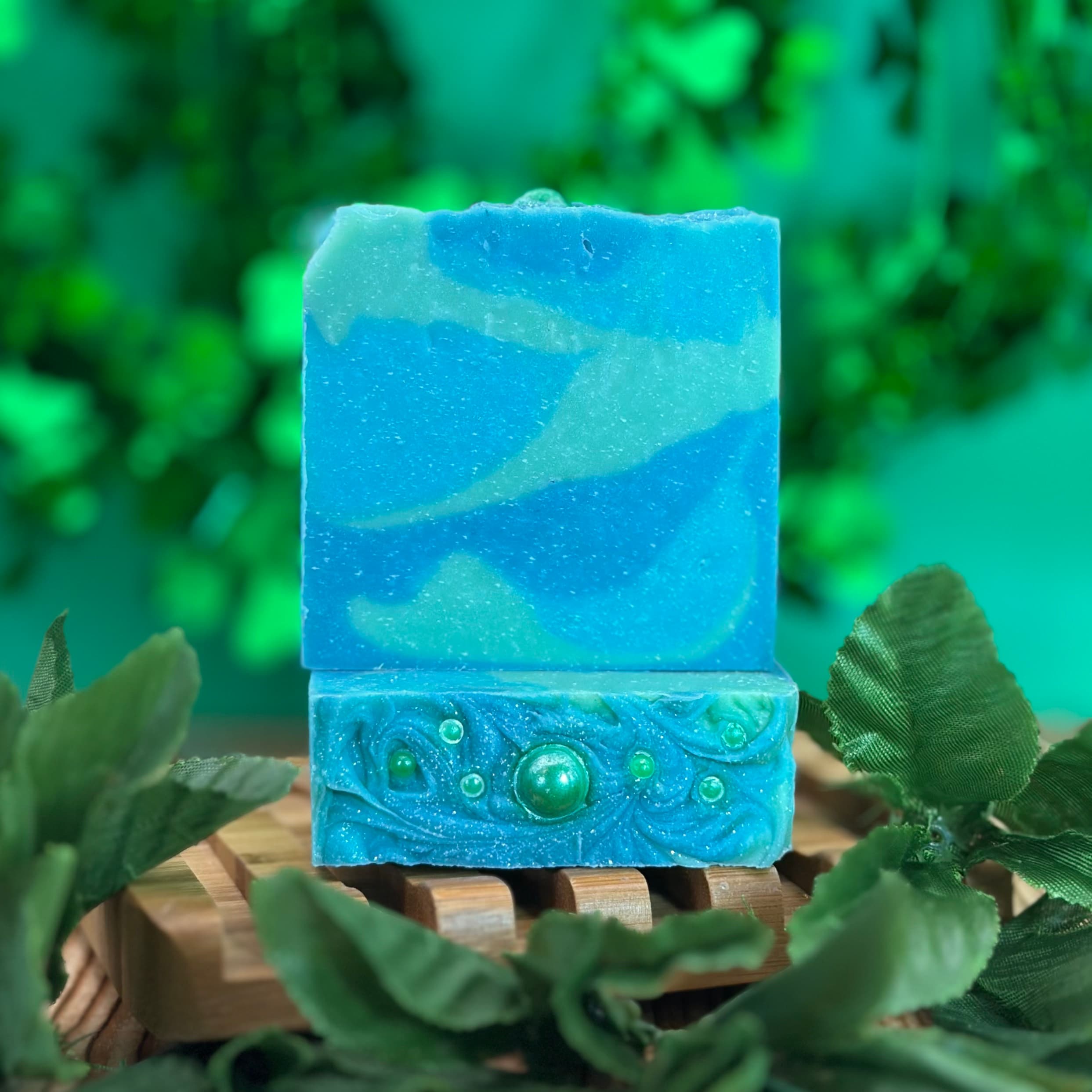 Two stacked blue and green marbled soap bars on a wooden soap dish. The bottom bar features decorative swirls with embedded green spherical embeds resembling bubbles or gems.