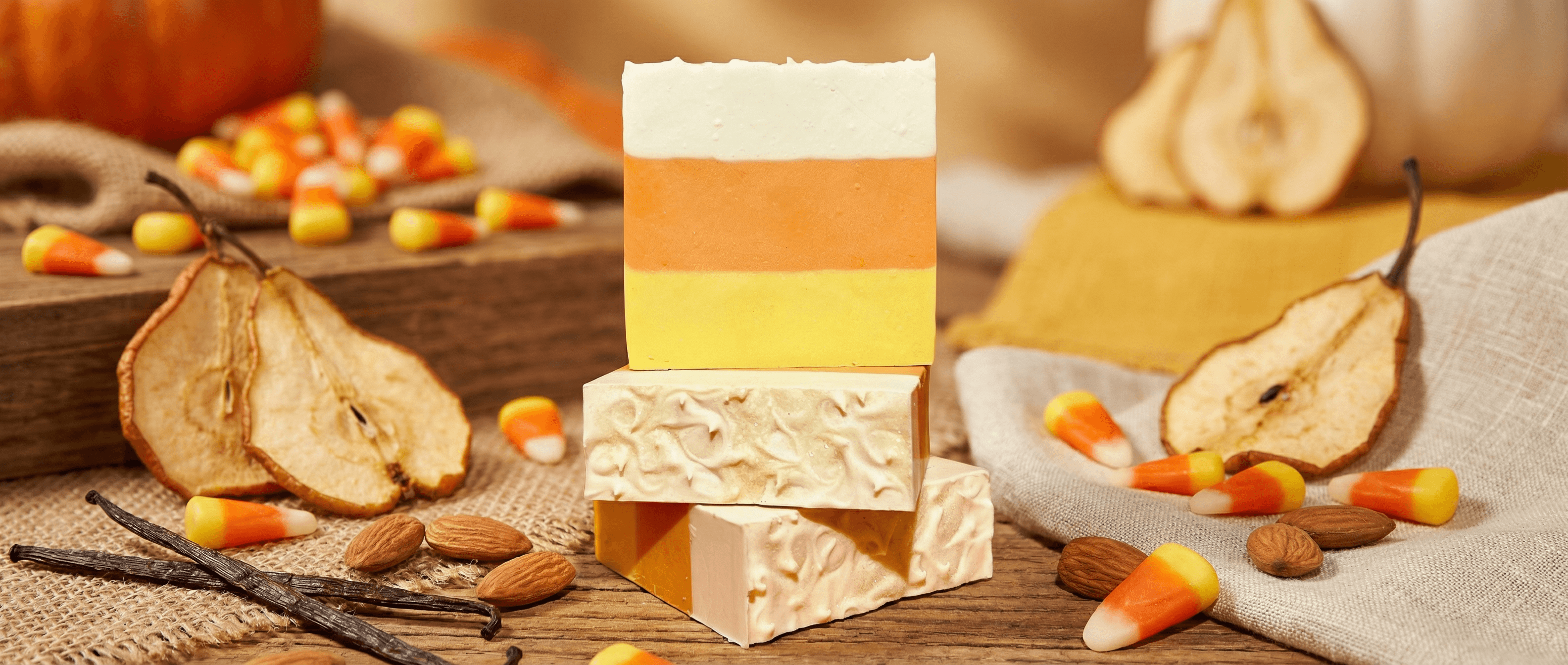 Handcrafted soap bars in candy corn colors: white top layer, orange middle, yellow bottom. Textured surface with oat flakes visible. Styled with dried pears, almonds, and vanilla beans.