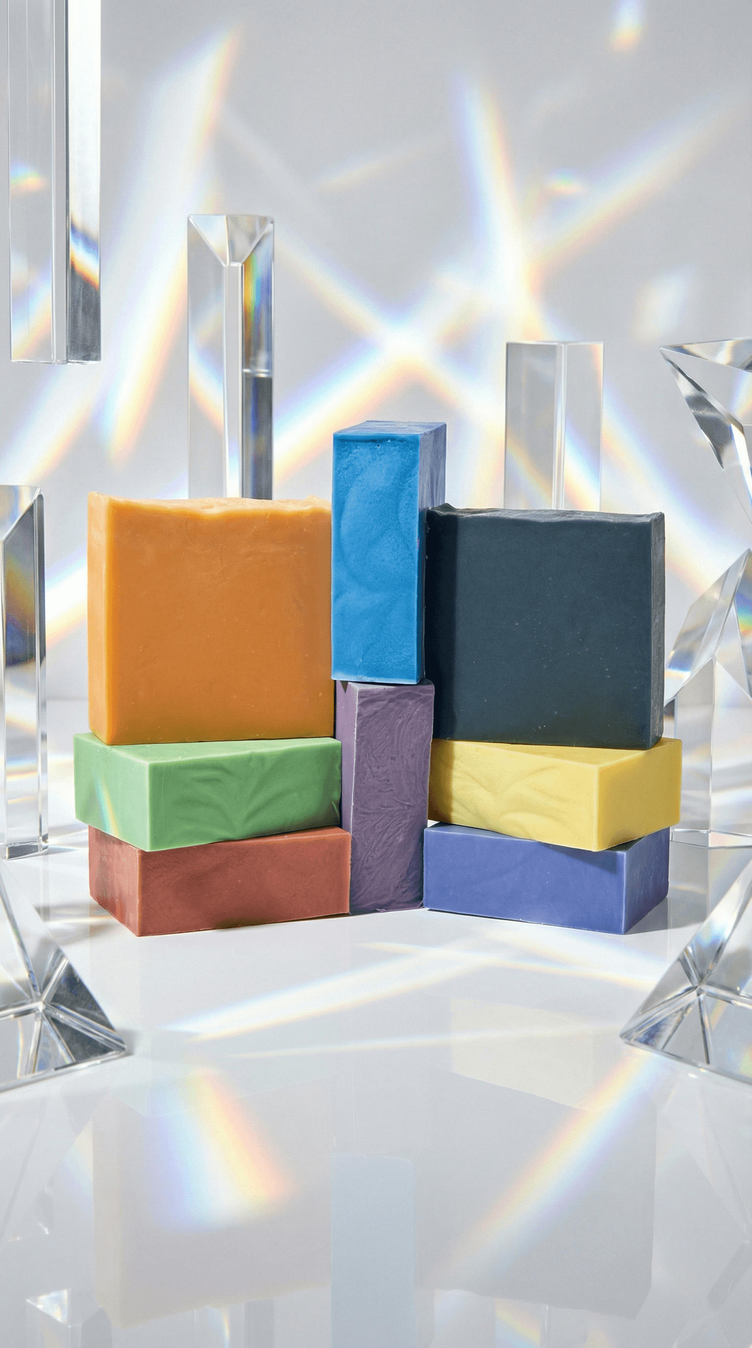 Eight colorful rectangular soap bars stacked together in rainbow order from red to violet plus black and yellow, surrounded by clear prisms casting rainbow light patterns.