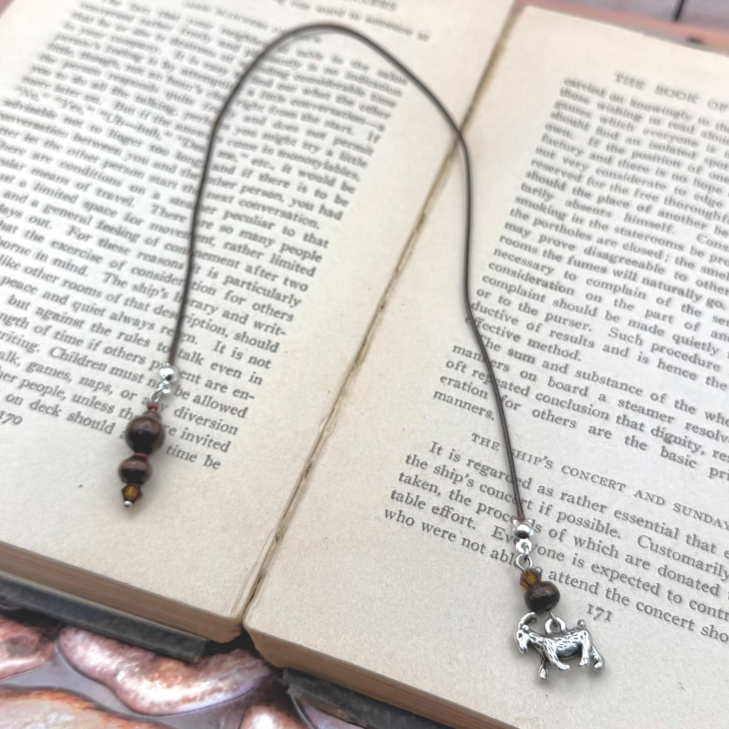 Handmade bookmark with black cord draped across open book pages. Features dark wooden beads and silver goat charm dangling at bottom. Natural, rustic styling with vintage book background.
