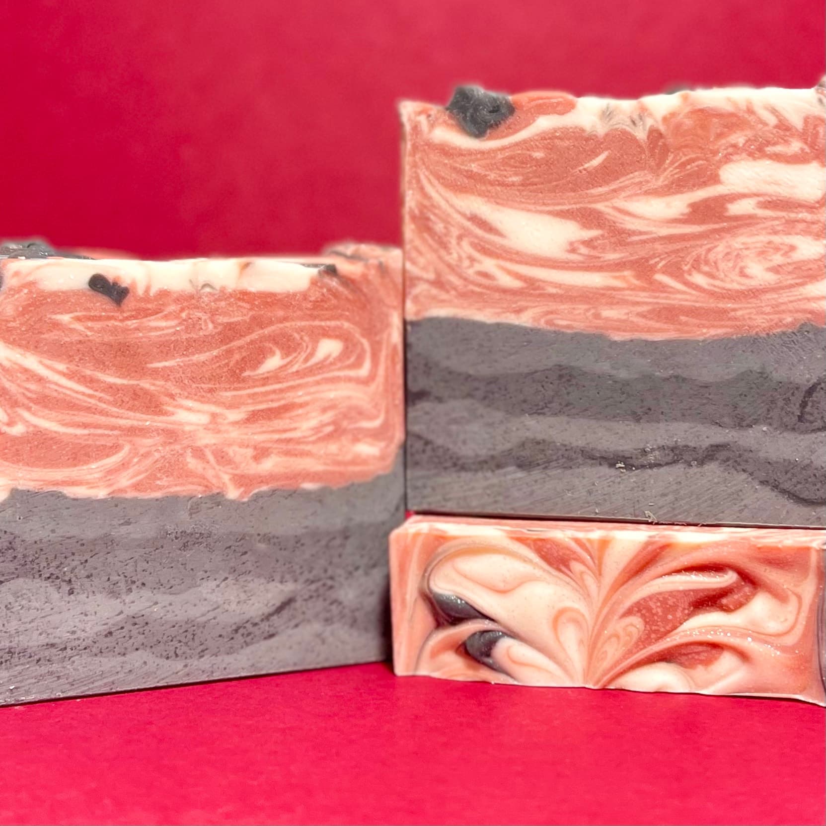 Three bars of handmade soap with swirled coral and cream tops, charcoal gray bases, and dark botanical inclusions on top, displayed against a bright pink background.