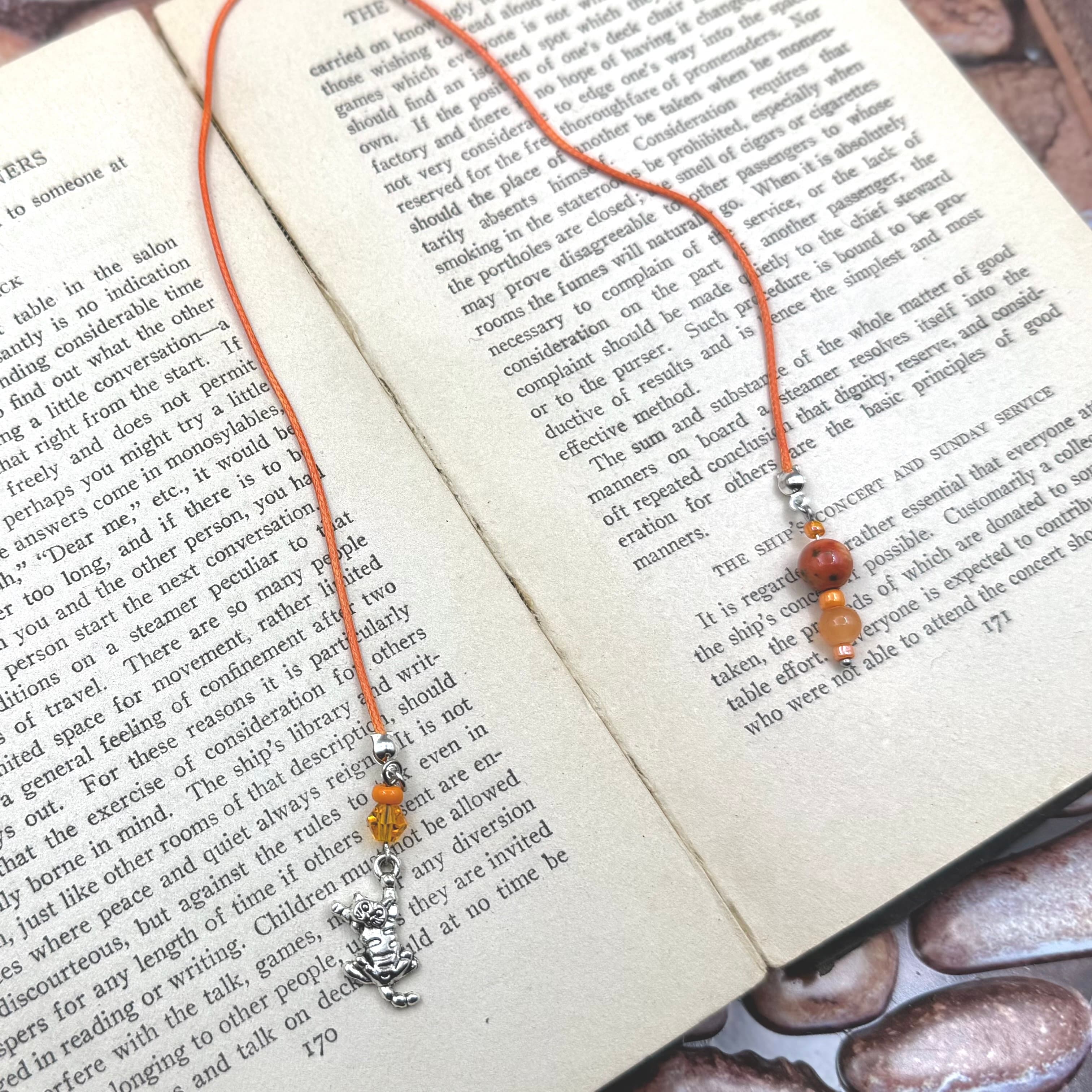 Orange cord bookmark with small beaded accents and silver cat charm, displayed on open book pages. Handmade design features warm amber and brown glass beads above decorative feline pendant.