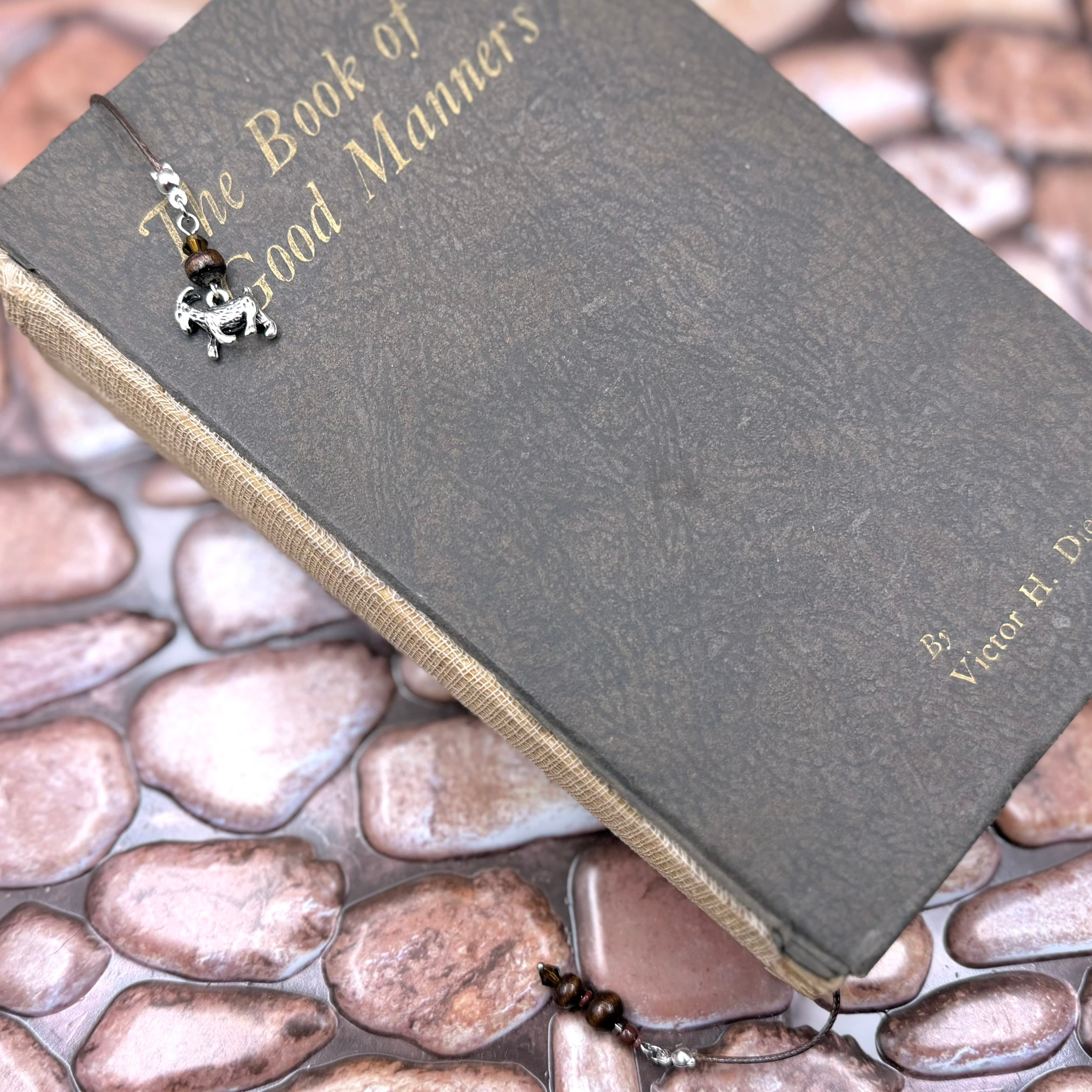 Gray hardcover book titled 'The Book of Good Manners' with tan cloth spine, resting on decorative stone surface. Silver goat charm bookmark with black and clear beads attached.