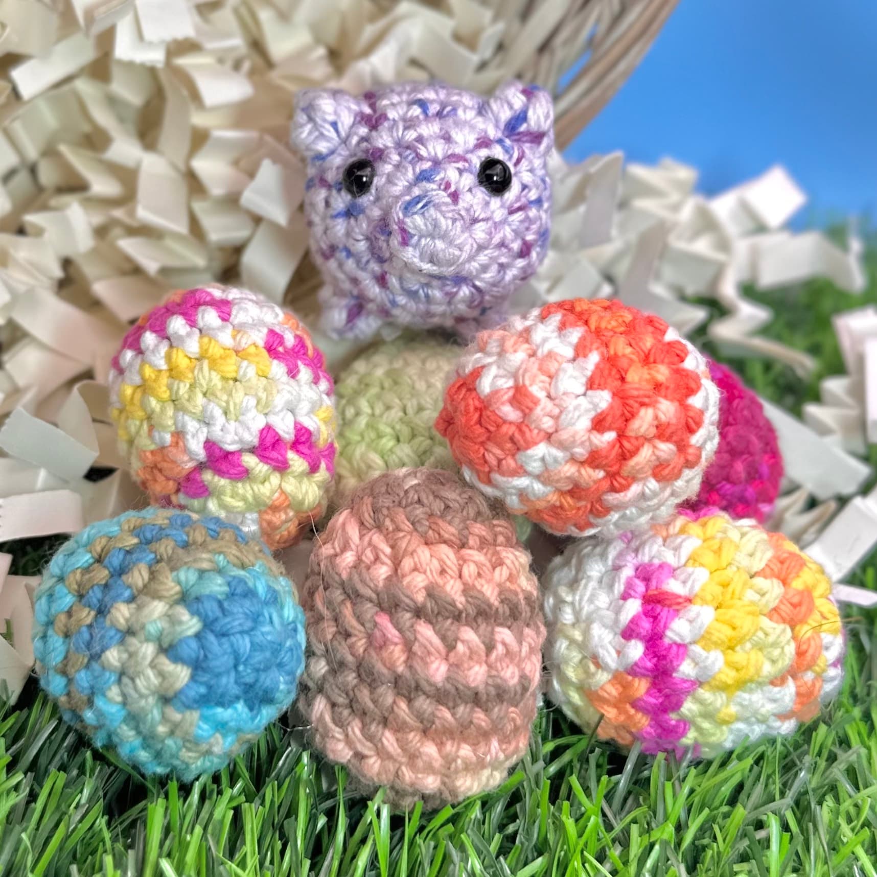 Seven colorful crocheted eggs in striped and solid patterns arranged on grass with white paper shred, topped by a small purple crocheted pig with black bead eyes.