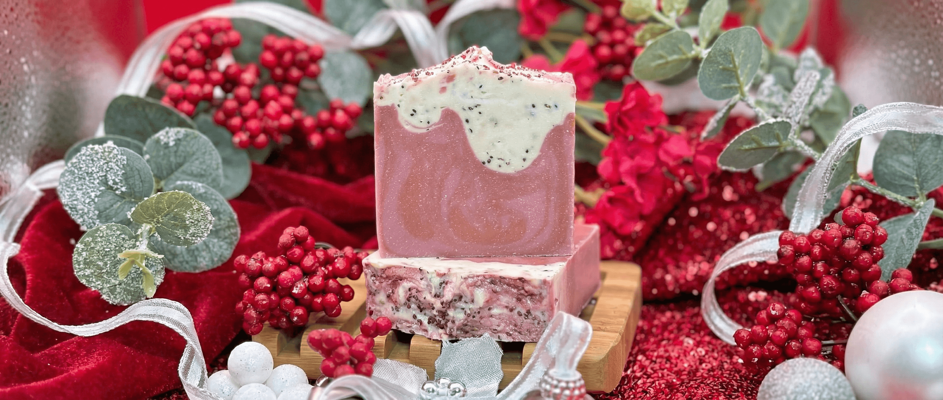 Pink handcrafted soap bar with white frosted top decorated with black and red speckles, displayed on wooden soap dish surrounded by red berries, eucalyptus leaves, and silver ribbons.