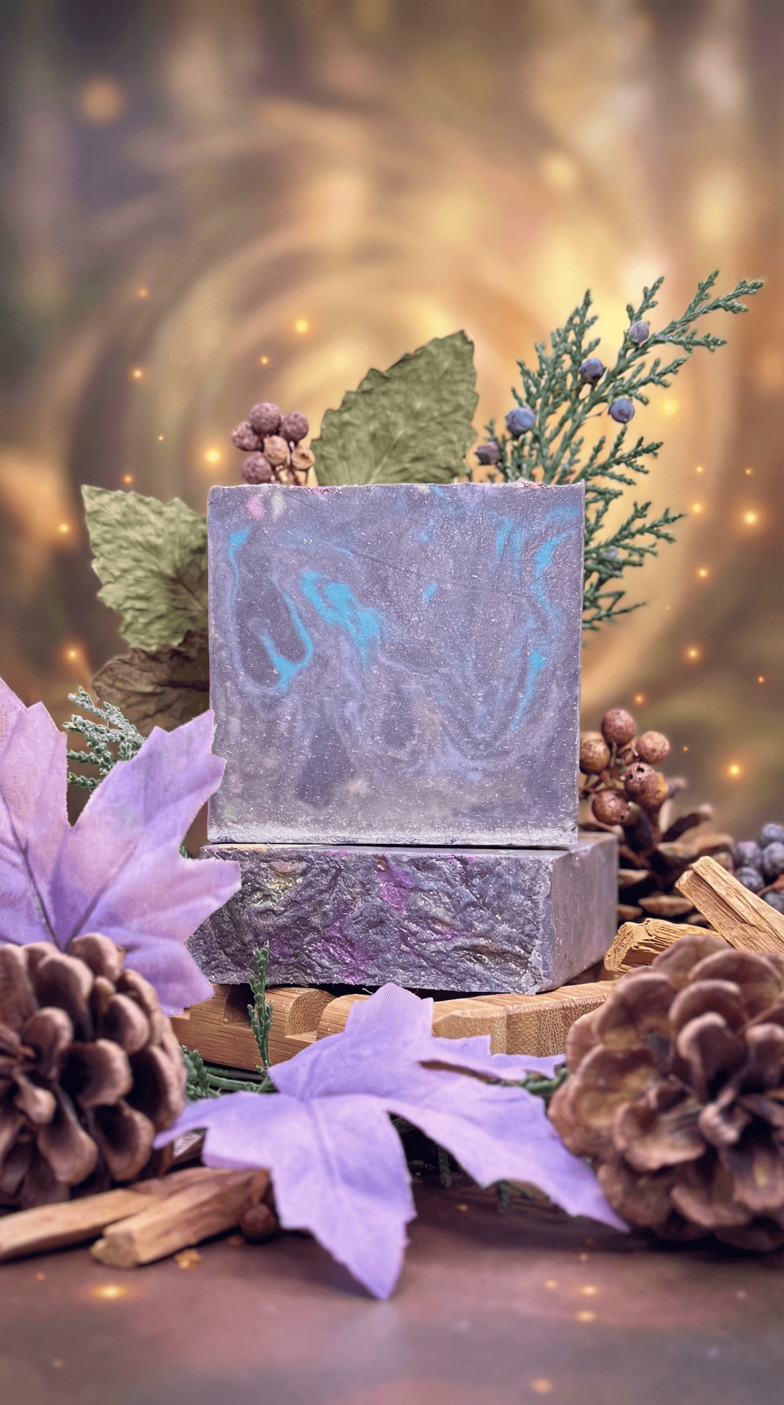 Two stacked purple and blue swirled soap bars with marbled texture, displayed among pine cones, dried botanicals, lavender flowers, and evergreen sprigs with warm lights in background.