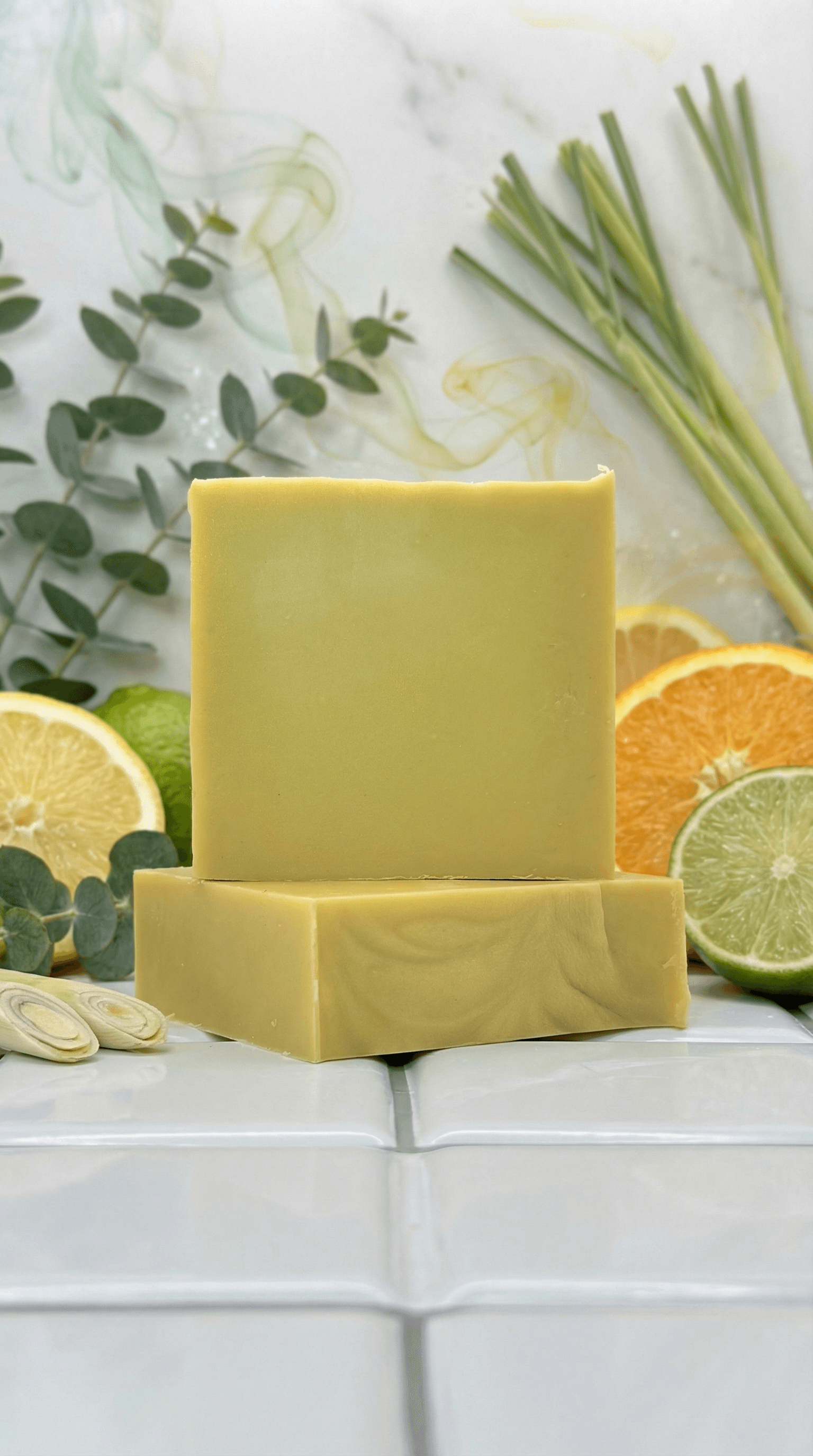 Pale yellow rectangular soap bars stacked on white tile, surrounded by fresh citrus slices, lemongrass stalks, and eucalyptus sprigs. Smooth matte surface with clean geometric edges.