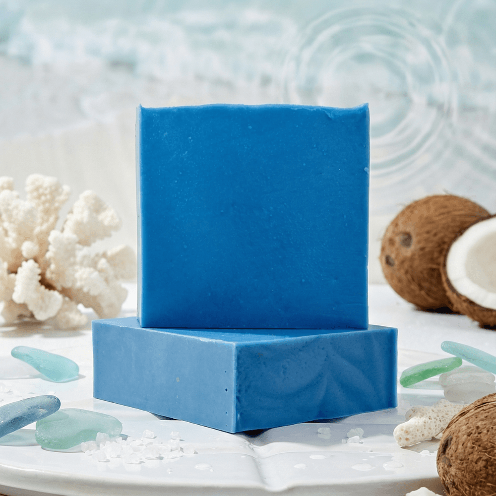Two bright blue rectangular soap bars stacked on a white marble surface, surrounded by white coral, coconut halves, sea glass, and seashells in an ocean-themed display.