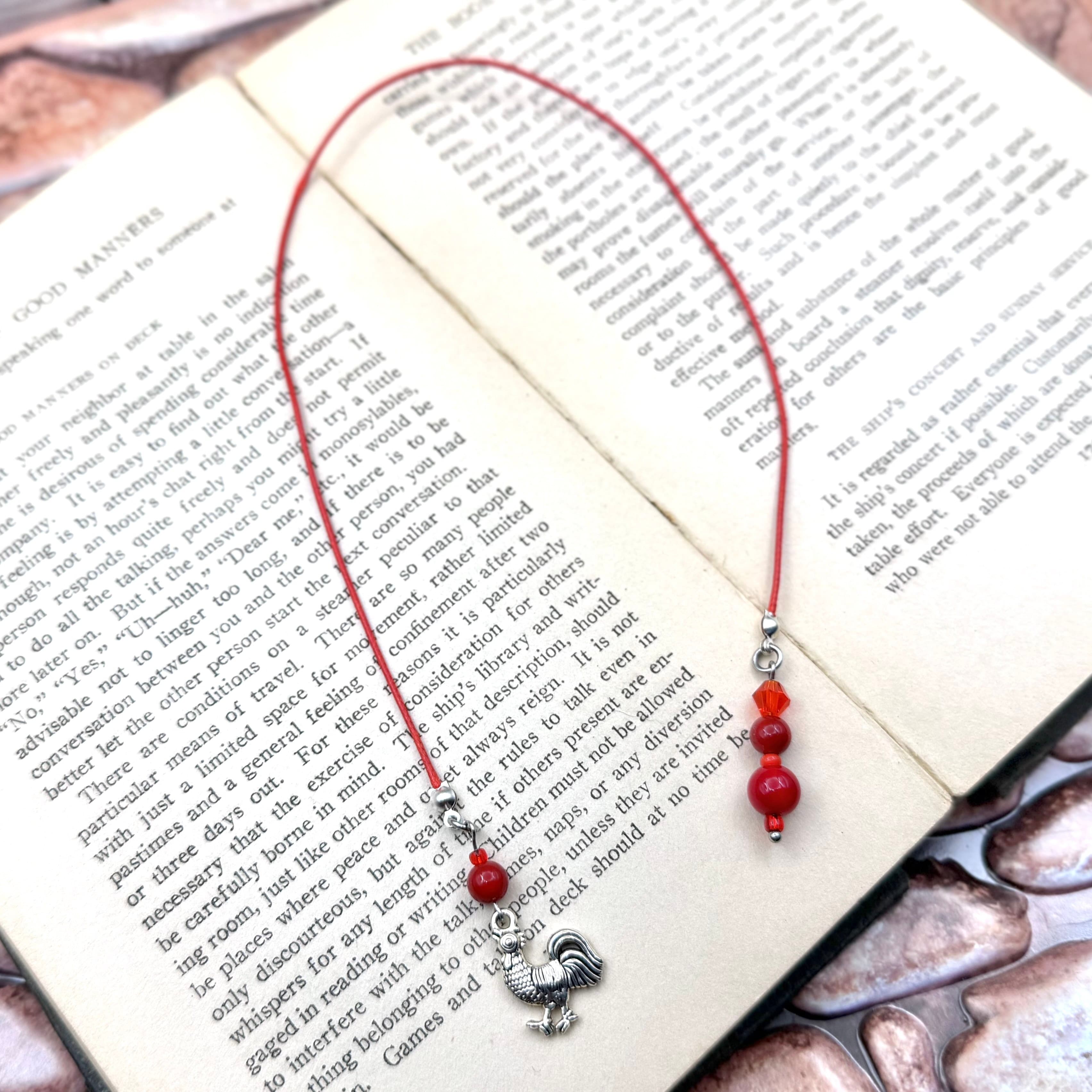 Handmade bookmark with red cotton cord forming a loop, decorated with red glass beads and a silver rooster charm, displayed on an open book with visible text.