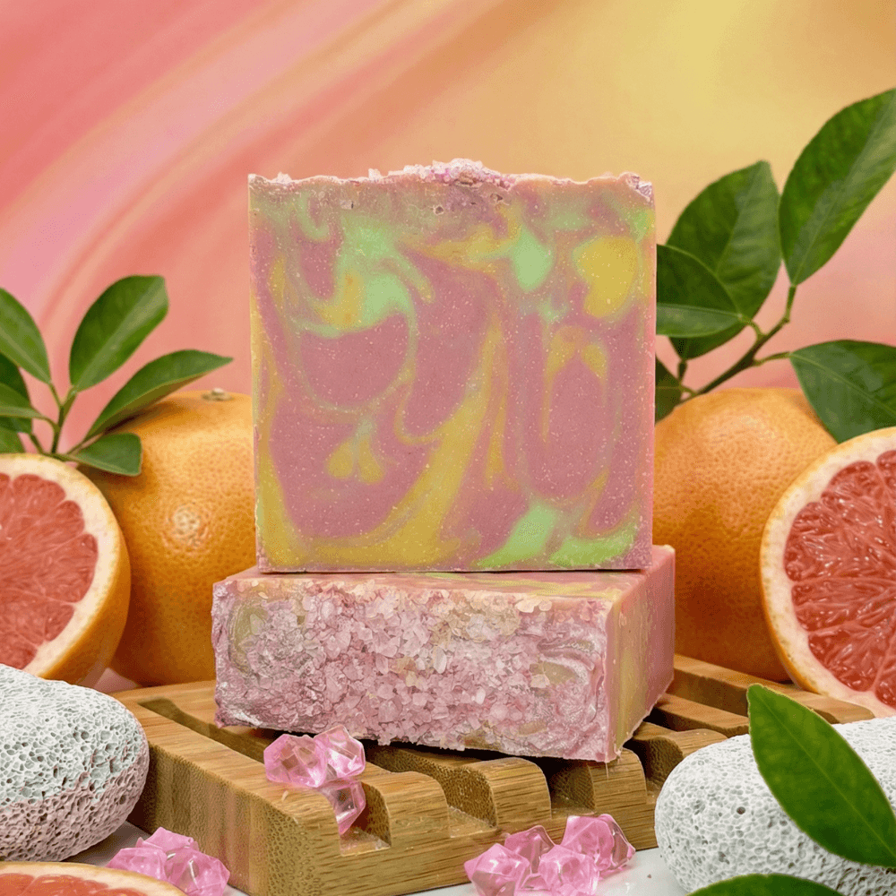 Two bars of swirled pink, yellow, and green soap stacked on bamboo, surrounded by grapefruits, green leaves, pumice stones, and pink salt crystals against a gradient background.