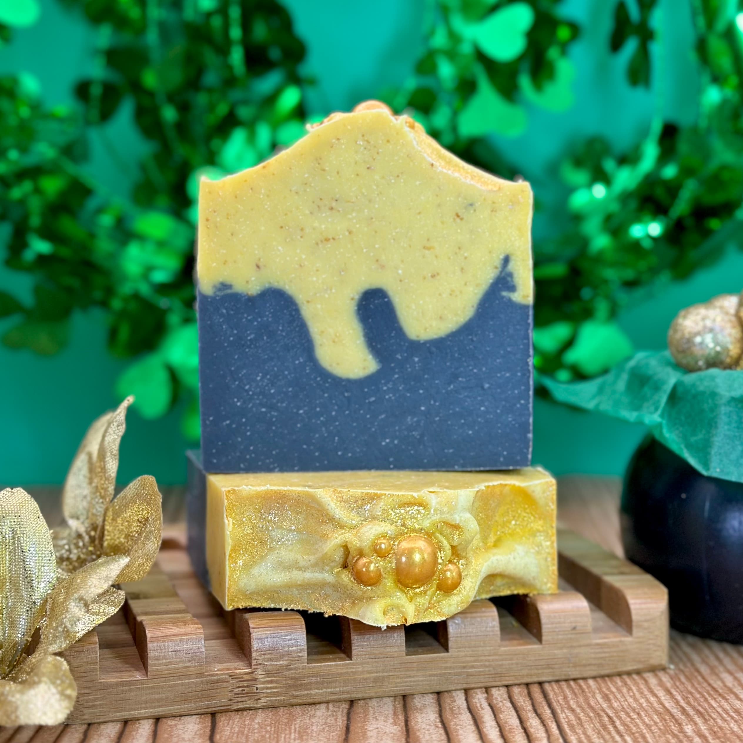 Two stacked handmade soap bars on a wooden soap dish with gold decorative leaves. Top bar features blue base with yellow dripping design, bottom bar is marbled gold with pearl embeds.