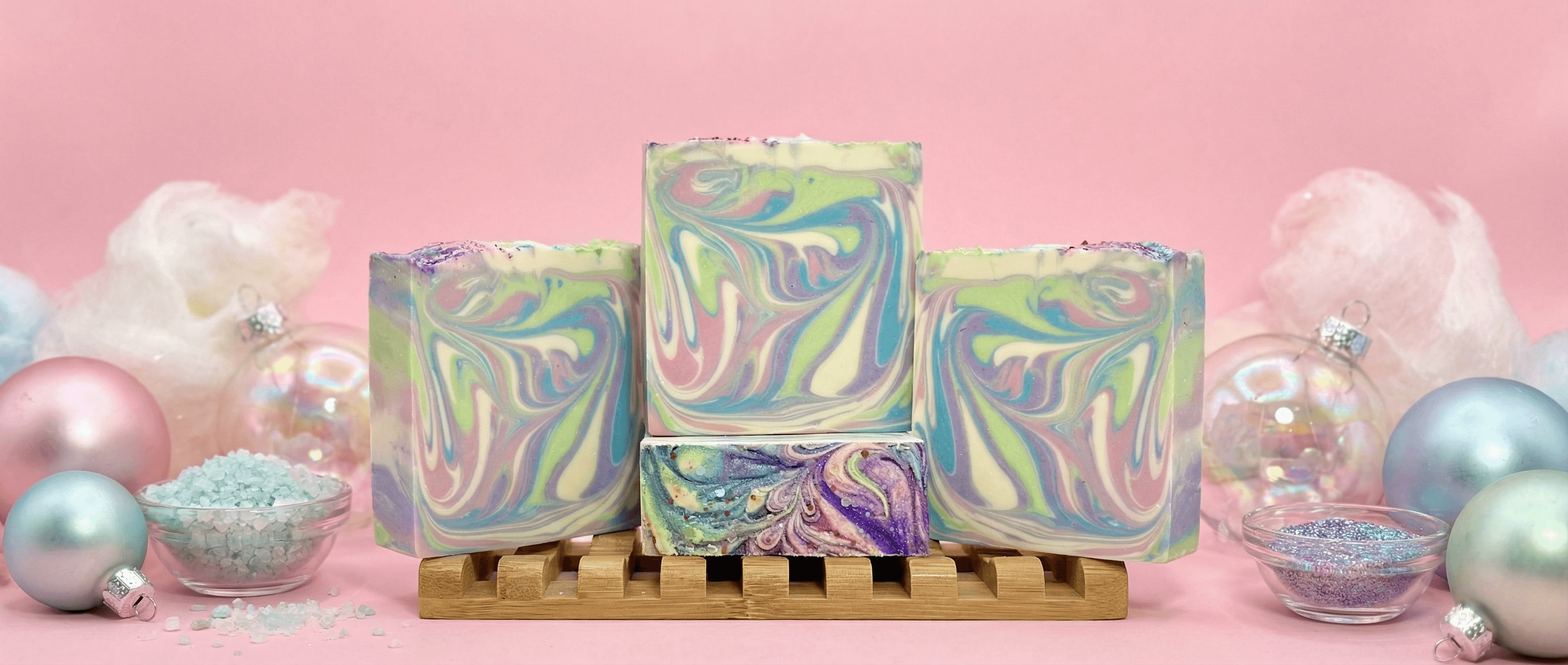 Four bars of handmade soap with swirled pastel green, yellow, pink, and blue marbling displayed on a wooden soap rack against a pink background with ornaments and bowls of glitter.