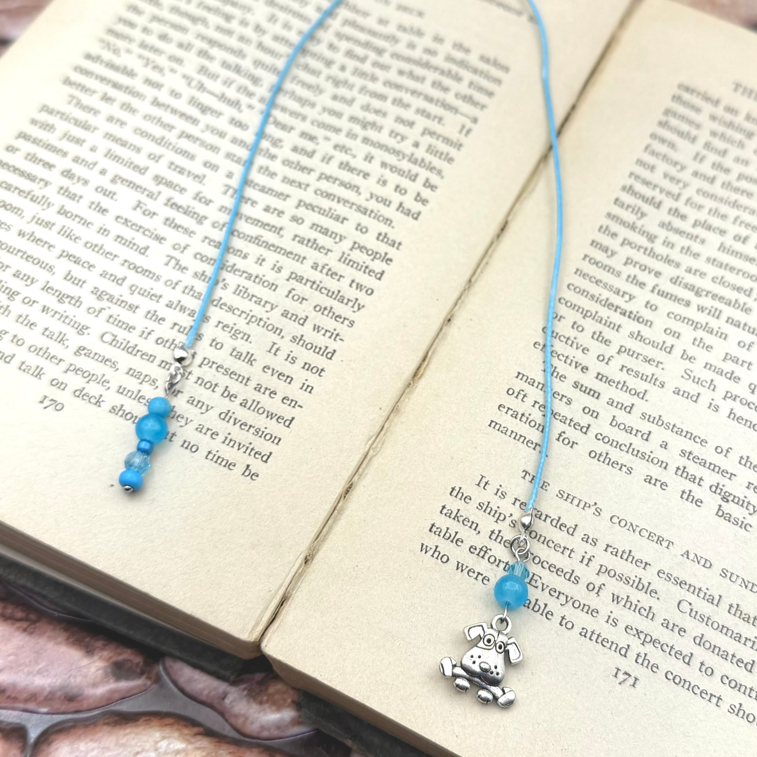 Handmade bookmark with blue cotton cord, light blue beads, and silver dog charm resting in open book showing printed text pages on cream-colored paper.