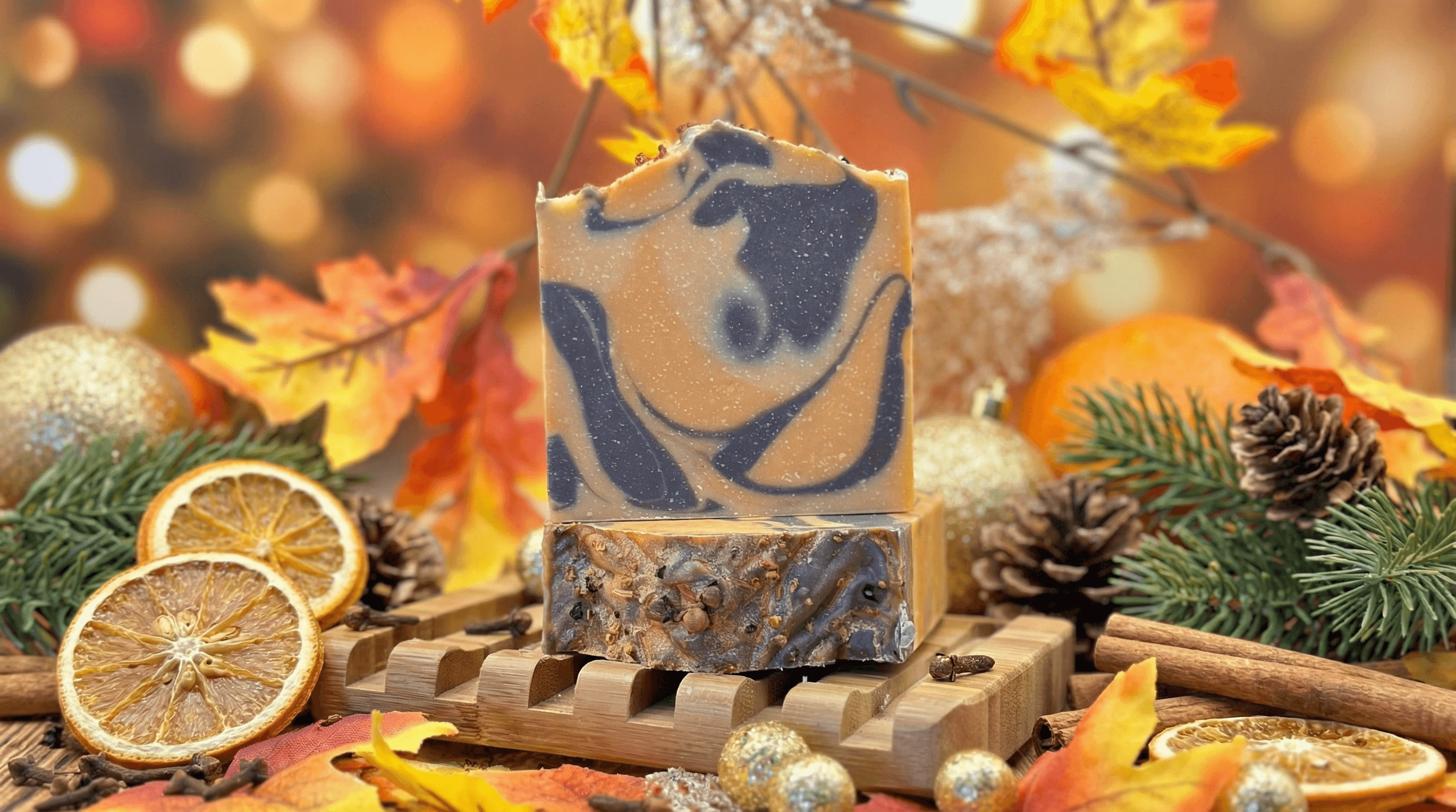 Two stacked bars of swirled tan and dark brown soap with marbled pattern, displayed on wooden soap dish surrounded by dried orange slices, pine cones, cinnamon sticks, and autumn leaves.
